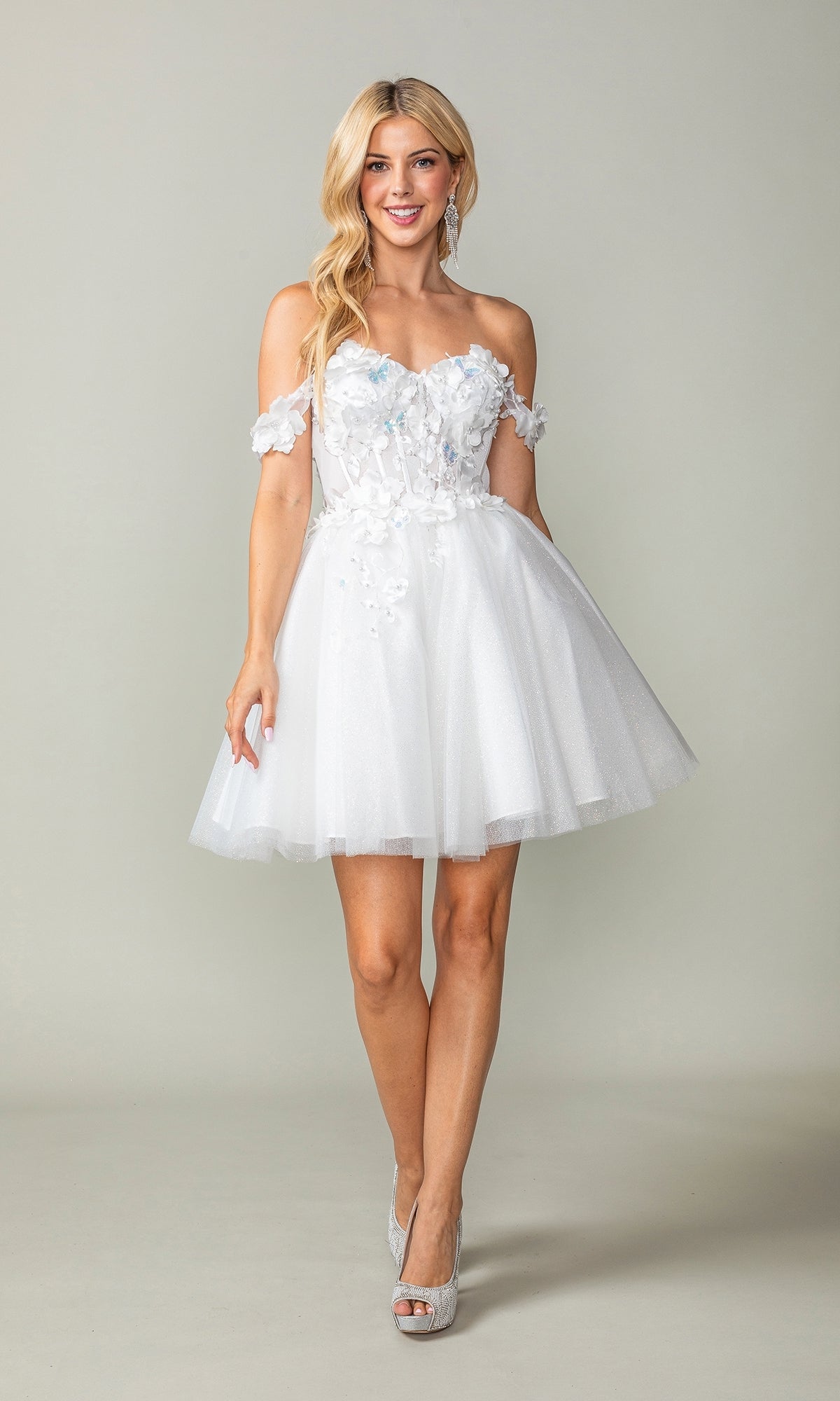 A woman poses in the Off-Shoulder Short Babydoll Homecoming Dress 3388, featuring floral appliqués and a tulle skirt, paired with silver heels. The short cocktail dress stands out beautifully against the plain background.
